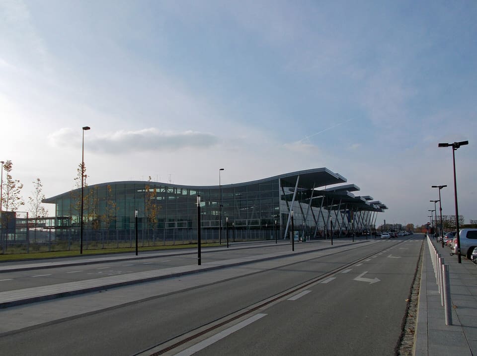 The glass front of Wrocław Airport's passenger terminal under its long wave-shaped roof on slanting steel columns, seen down the empty approach road on an autumn afternoon.