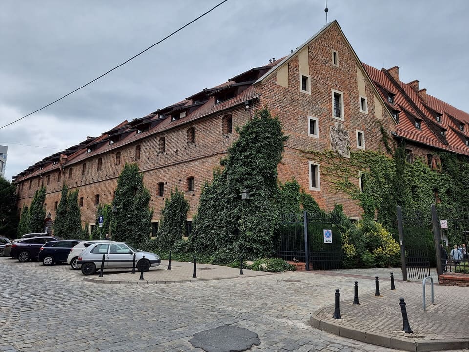 The long brick facade of the City Arsenal, part of it under ivy, with a red tiled roof, seen from the street corner.