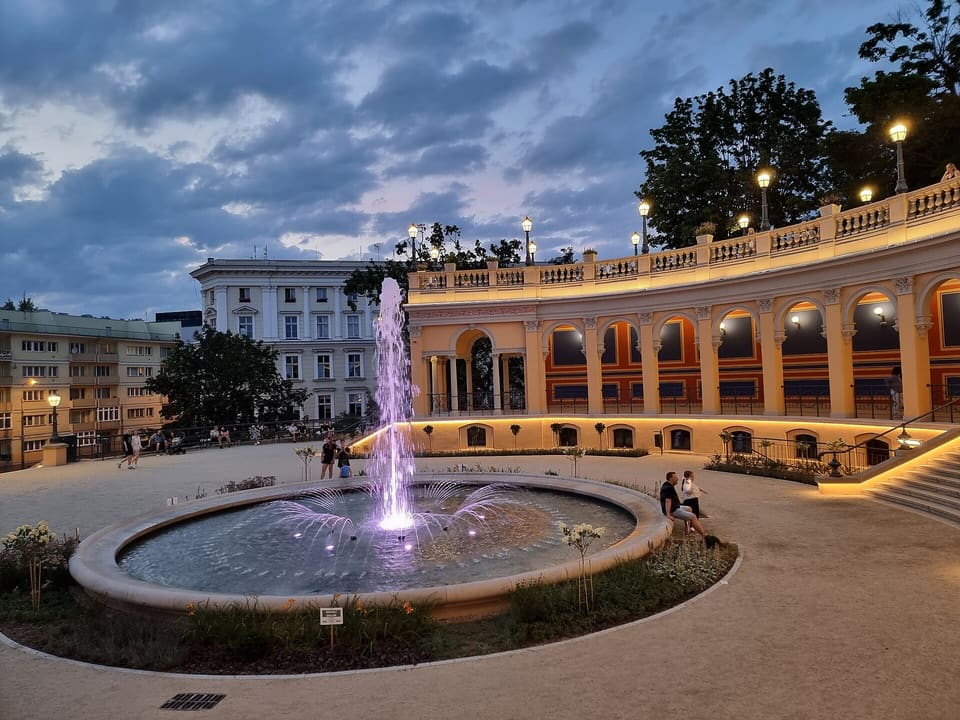 The curved colonnade of the Bastion Sakwowy at dusk, lit from below, with the round fountain in the square in front of it.