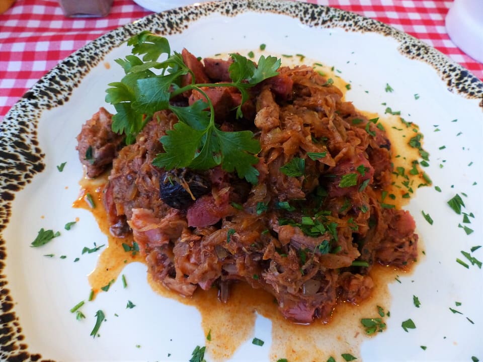 Ein Teller Bigos: geschmortes Sauerkraut und Kohl mit Fleisch- und Wurststücken, bestreut mit gehackter Petersilie und gekrönt von einem Petersilienzweig, auf einem weißen Teller mit gesprenkeltem Rand.
