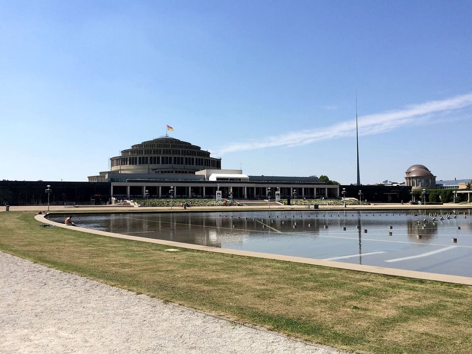 The stepped round dome of Centennial Hall and its long colonnaded front seen across the still water of the fountain's pool, with the steel spire of the Iglica to the right, under a clear sky.