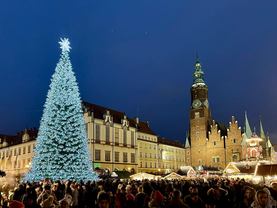 Ein großer beleuchteter Weihnachtsbaum und Marktstände auf dem Marktplatz in der Dämmerung, im Vordergrund eine Menschenmenge, dahinter der gotische Uhrturm des Alten Rathauses.