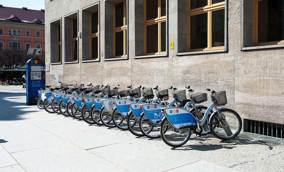 Eine Reihe blau-weißer Leihfahrräder in ihren Halterungen entlang einer steinernen Hauswand, am Ende das Terminal der Station.