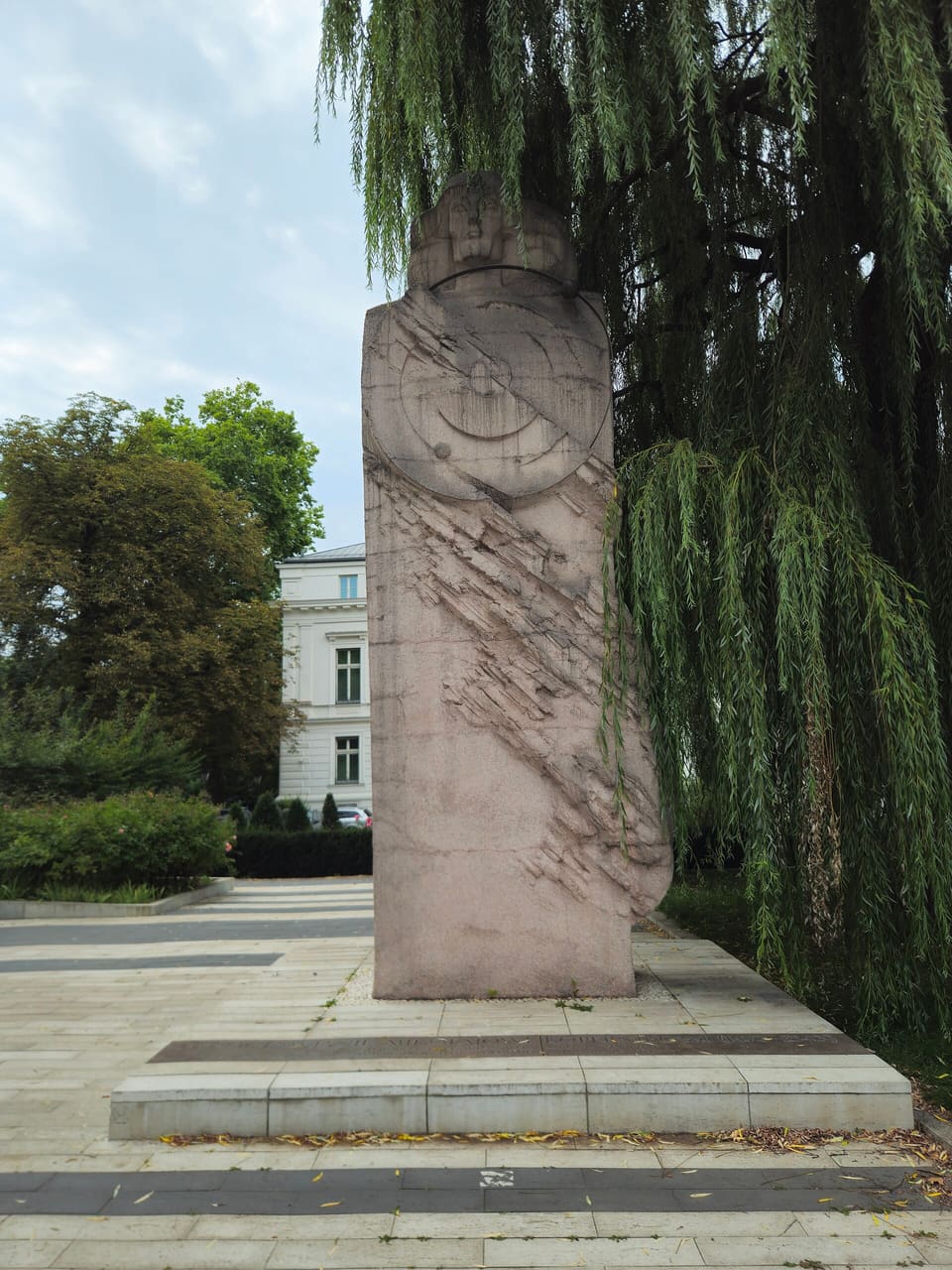 The Copernicus monument: a tapering stone stele carved with a disc of planetary orbits and topped by a stylised head, on a stepped paved base beside a willow.