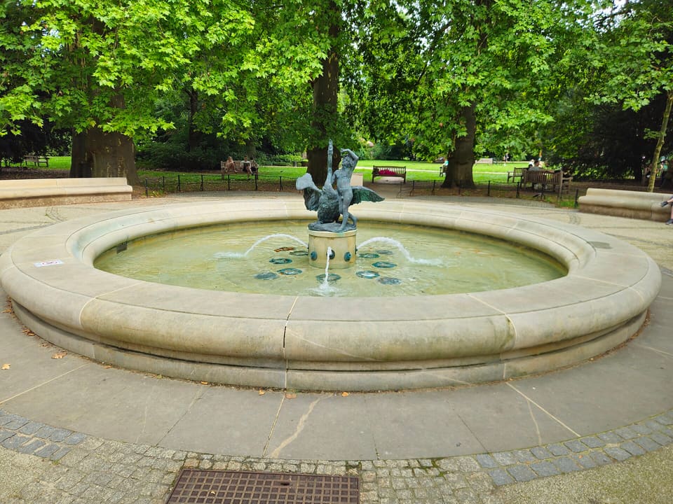 A circular stone fountain in the park, with the bronze figure of Cupid on Pegasus above its basin, among mature trees and lawns.