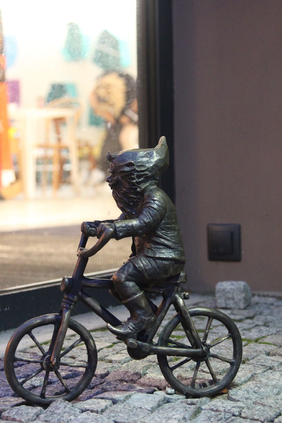 Eine kleine Zwergfigur aus Bronze, die auf einem Fahrrad eine gepflasterte Straße entlangfährt und mitten im Tritt den Lenker umfasst.