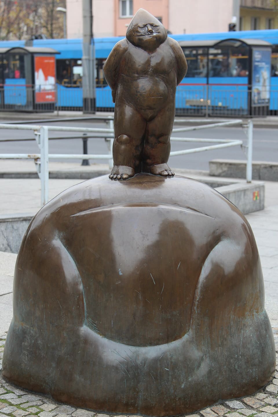 Eine größere Zwergfigur aus Bronze mit den Händen auf dem Rücken, auf einem abgerundeten Felsblock als Sockel an der Straße, dahinter eine Straßenbahn und ein Geländer.