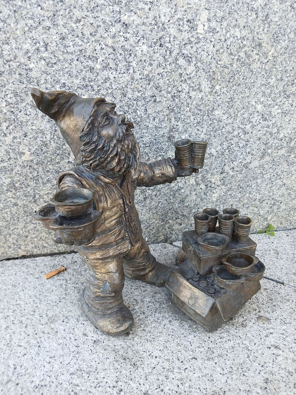 Eine kleine Zwergfigur aus Bronze, die in jeder Hand gestapelte Sammelbüchsen hält, auf einem Steinpflaster vor einer Granitwand.
