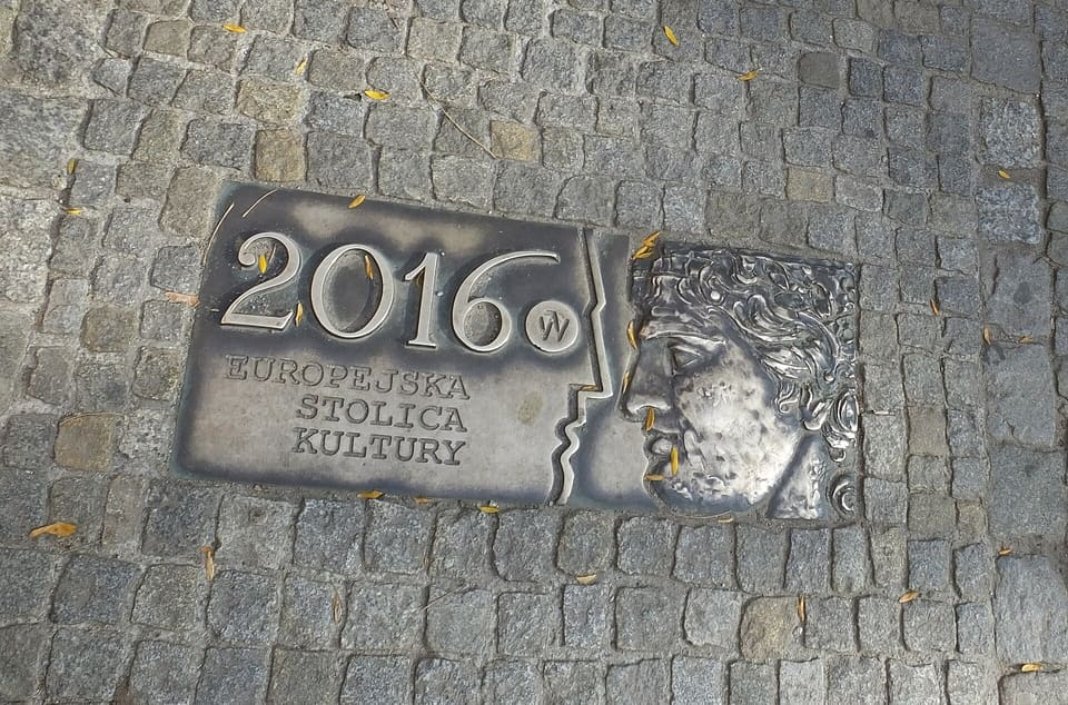 A bronze plaque set into granite cobbles, lettered 2016 EUROPEJSKA STOLICA KULTURY beside a face in relief.