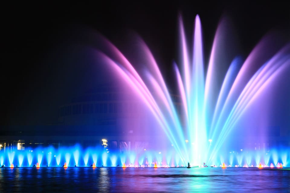 Jets of the fountain lit in blue, purple and green against a dark sky, with the curved dome of the Centennial Hall behind them.