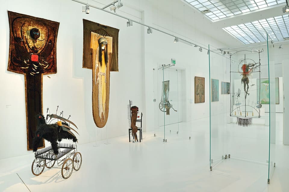A white top-lit gallery inside the pavilion hung with large textile works, a black feathered figure in a pram on the floor and glass display cases along the right-hand wall.