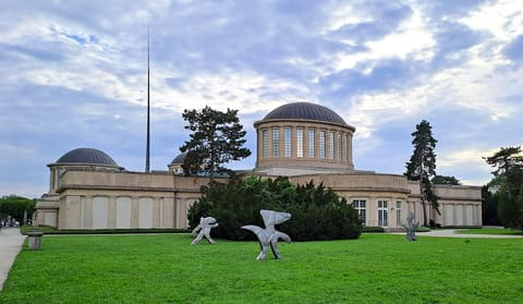 Helle Skulpturen auf dem Rasen vor dem Pavillon, dahinter eine Kuppel auf ihrem Tambour und das lange, niedrige Gebäude, in der Ferne eine schlanke Spitze.