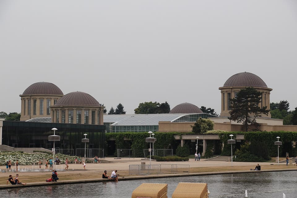 The Four Domes Pavilion seen across the fountain pool: four low domes on colonnaded drums above a long glazed hall, with people sitting at the water in front of it.