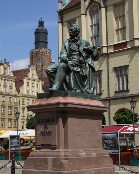 Die sitzende Bronzestatue von Aleksander Fredro auf ihrem Sandsteinsockel mit der Inschrift ALEKSANDER HR. FREDRO, in der Nachmittagssonne auf dem Marktplatz, dahinter der Turm der Kirche St. Elisabeth und barocke Giebel.