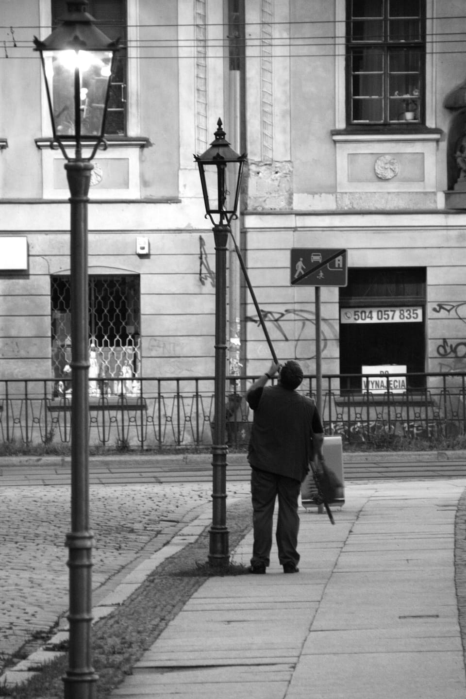 A black and white night photograph of a lamplighter seen from behind, reaching up with a long pole to light a street lamp, with a second lamp already burning beside him.