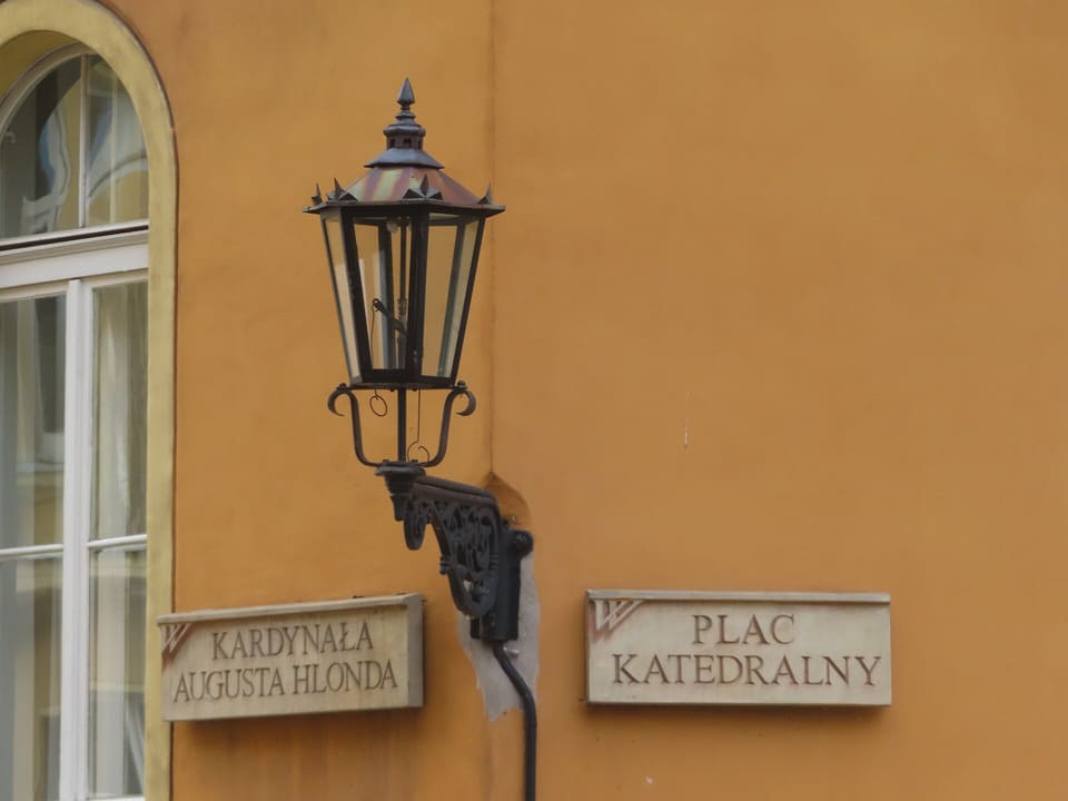Eine schwarze schmiedeeiserne Gaswandlaterne mit kupferfarbenem Dach an einer ockerfarbenen Wand, neben zwei steinernen Straßenschildern mit den Aufschriften Kardynała Augusta Hlonda und Plac Katedralny.