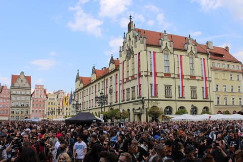 Eine dichte Menge mit Gitarren füllt den Marktplatz vor dem gelben neugotischen Neuen Rathaus, dessen Fassade mit senkrechten rot-weißen Fahnen behängt ist, links die Giebelhäuser des Platzes unter blauem Himmel.