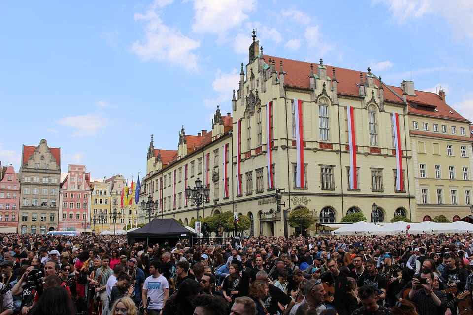 A dense crowd holding guitars fills the Market Square in front of the yellow neo-Gothic New Town Hall, its facade hung with vertical red-and-white flags, the square's gabled houses to the left under a blue sky.