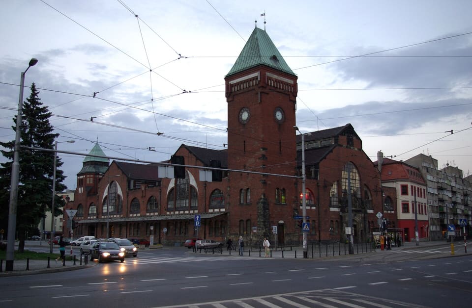 The Market Hall's red brick tower and curved gables seen from the street, with tram wires overhead.
