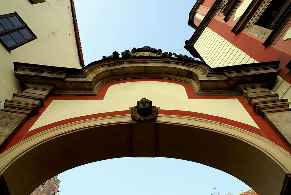 Nahansicht mit Blick nach oben durch den steinernen Torbogen, der die beiden Häuser verbindet, mit der gemeißelten Maske am Schlussstein und dem geschwungenen Gesims vor dem Himmel; durch die Öffnung darunter ist ein Turm zu sehen.