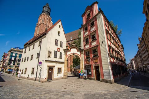 Zwei aneinandergrenzende historische Häuser, eines weiß mit steilem rotem Dach, eines rosa-weiß mit geschwungenem Giebel, verbunden durch einen Torbogen neben dem Backsteinturm der Kirche St. Elisabeth.