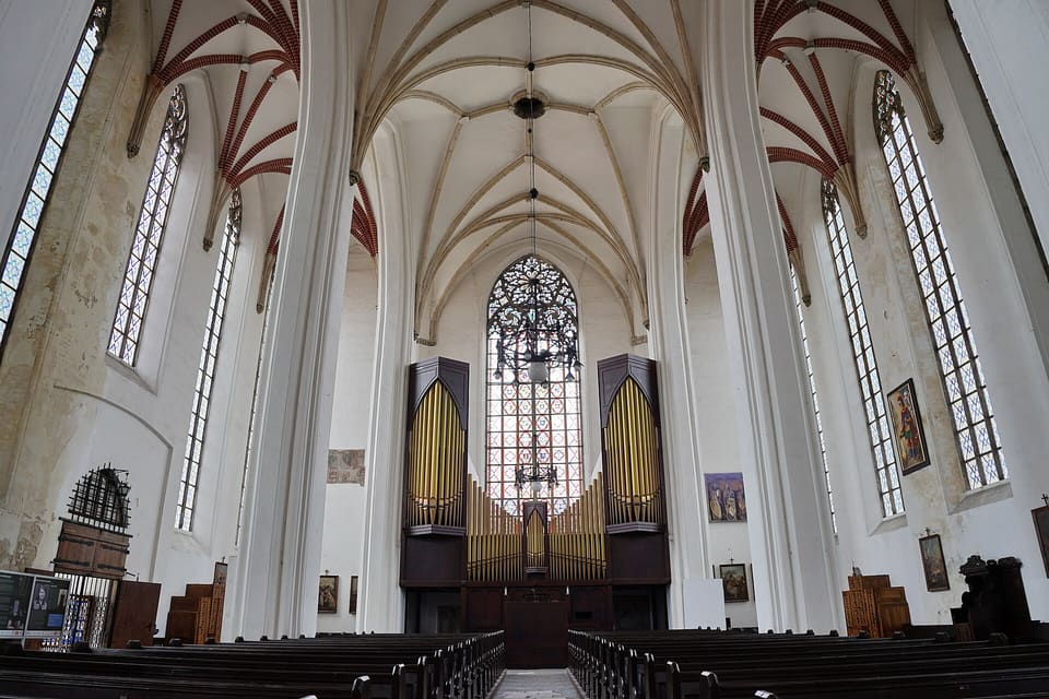 Looking down the nave of the upper church between tall white pillars to the organ with gilded pipes in front of a large west window, under a Gothic net vault with red-brick ribs in the side aisles and rows of dark pews below.