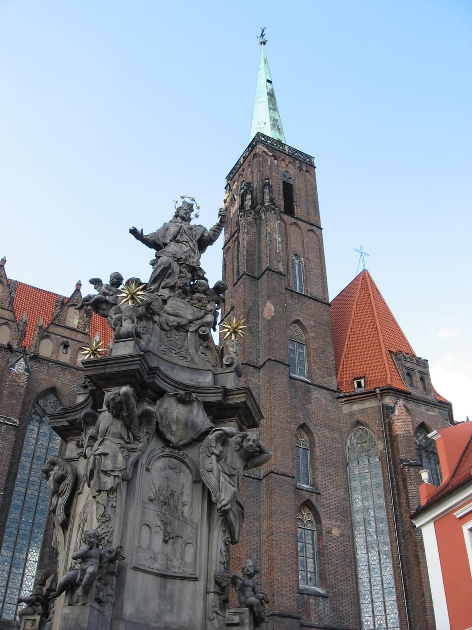 The brick tower of the church with its green copper spire, seen past the grey Baroque monument of St John of Nepomuk, which fills the lower left with a saint on top, angels and cherubs below and gilded stars.