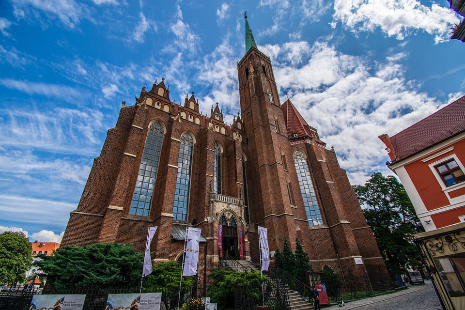 Looking up at the red-brick Gothic church from the street: tall pointed windows between buttresses, a gabled parapet along the roofline and a tall tower with a green spire behind, under a blue sky with white clouds.