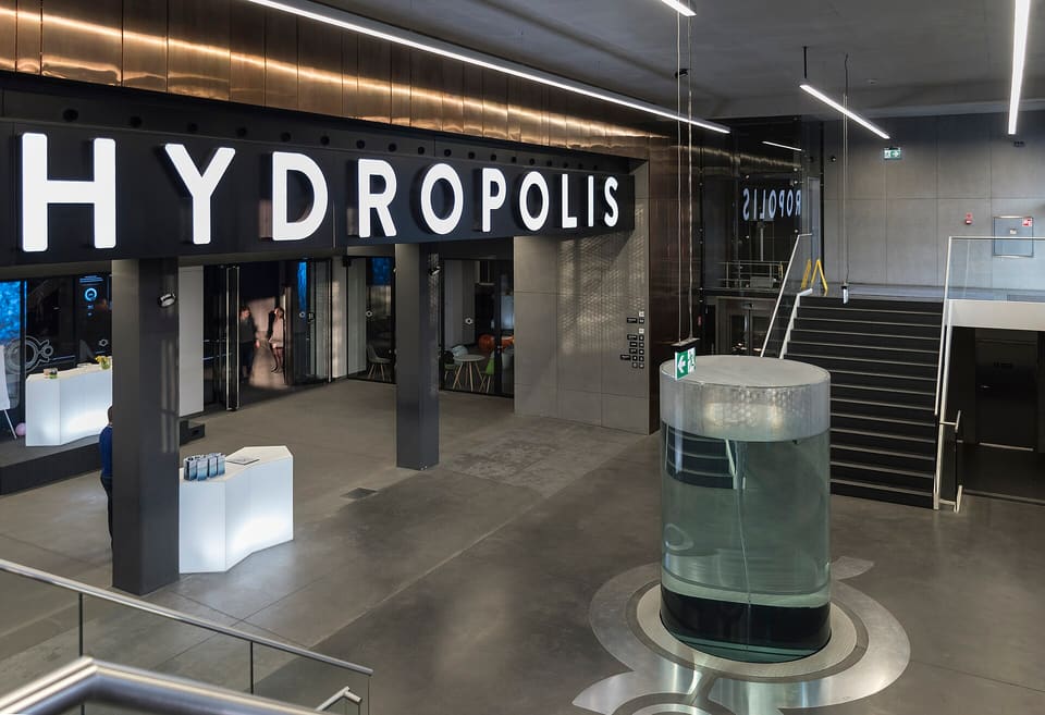 The foyer below ground: large illuminated HYDROPOLIS letters on a dark beam under a copper-clad ceiling, a tall glass cylinder of water standing on a steel floor medallion, and polished concrete underfoot.