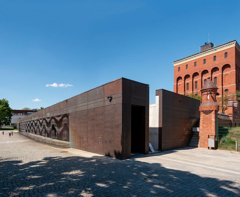 The long low entrance wall of weathered copper seen at an angle, with a perforated bay, a dark recessed doorway and the water-curtain rail above it, a cobbled forecourt in front and the red-brick arcaded water tower of the old waterworks rising behind.