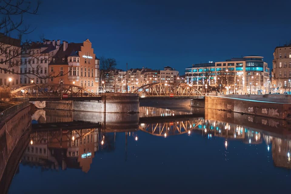 Zwei eiserne Bogenbrücken überqueren in der Abenddämmerung parallele Arme der Oder; ihre beleuchteten Bögen und die Fenster der umliegenden Gebäude spiegeln sich im stillen Wasser.