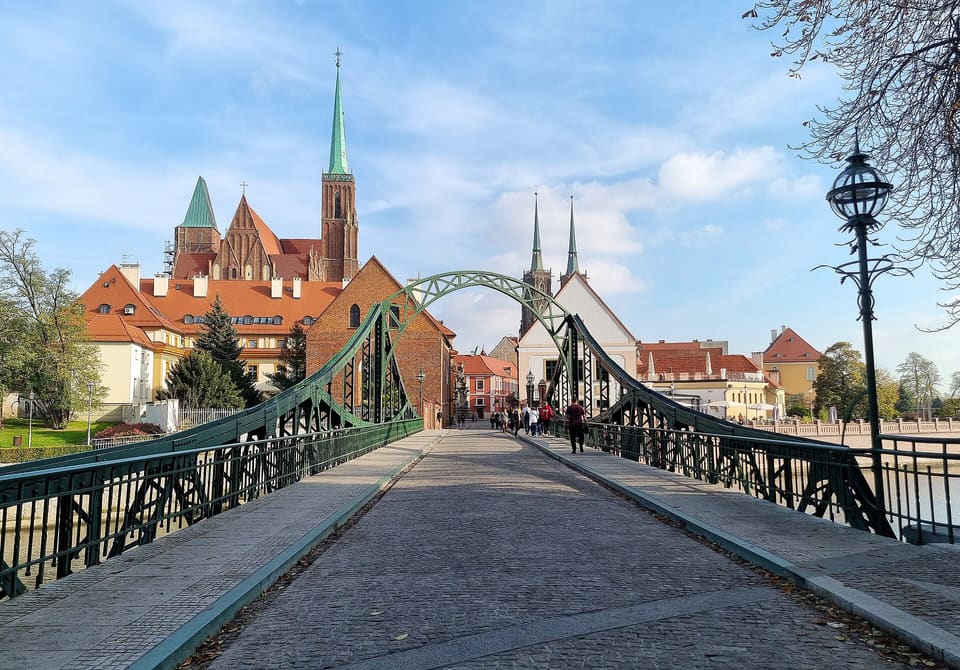 Eine gepflasterte Fußgängerbrücke mit grünem Eisenbogen führt auf die rot gedeckten Gebäude und Kirchtürme von Ostrów Tumski zu, unter einem teils bewölkten blauen Himmel, rechts eine Laterne und vorn einige Spaziergänger.