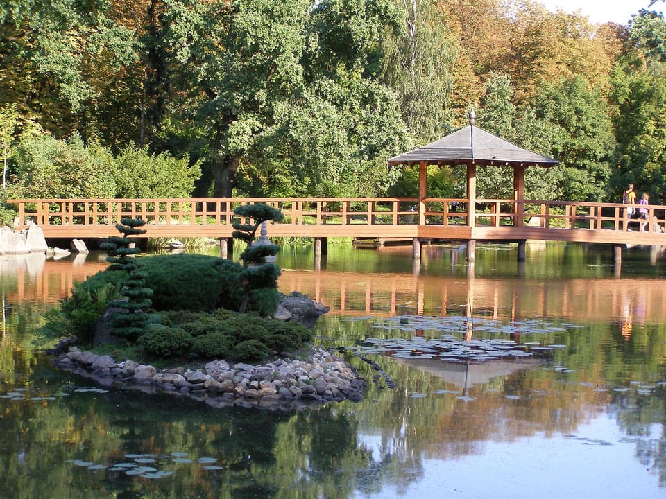 Eine kleine, von Steinen eingefasste Insel mit beschnittenen Kiefern in einem Teich, dahinter ein hölzerner Steg und ein Pavillon, im Hintergrund Bäume.