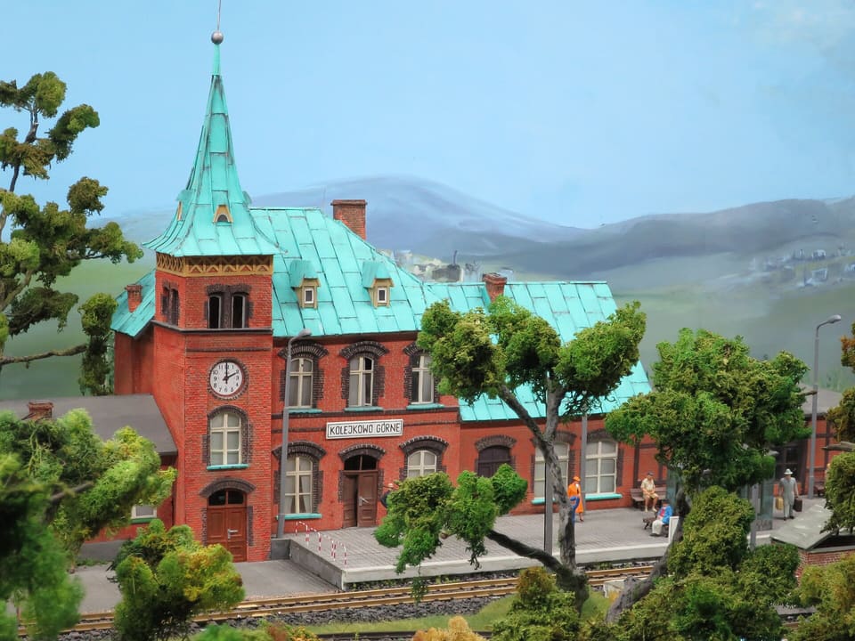 Eine Modelleisenbahnszene mit einem Bahnhofsgebäude aus rotem Backstein unter blaugrüner Turmspitze und blaugrünem Dach, vor gemalten Bergen, mit kleinen Figuren auf dem Bahnsteig.