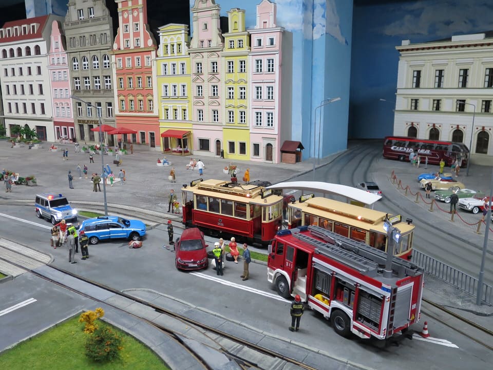 Eine Straßenszene im Modell mit einer Reihe bunter Miniatur-Mietshäuser, zwei Modellstraßenbahnen, einem Feuerwehrauto, Polizei- und Rettungsfiguren und einem gemalten blauen Hintergrund.
