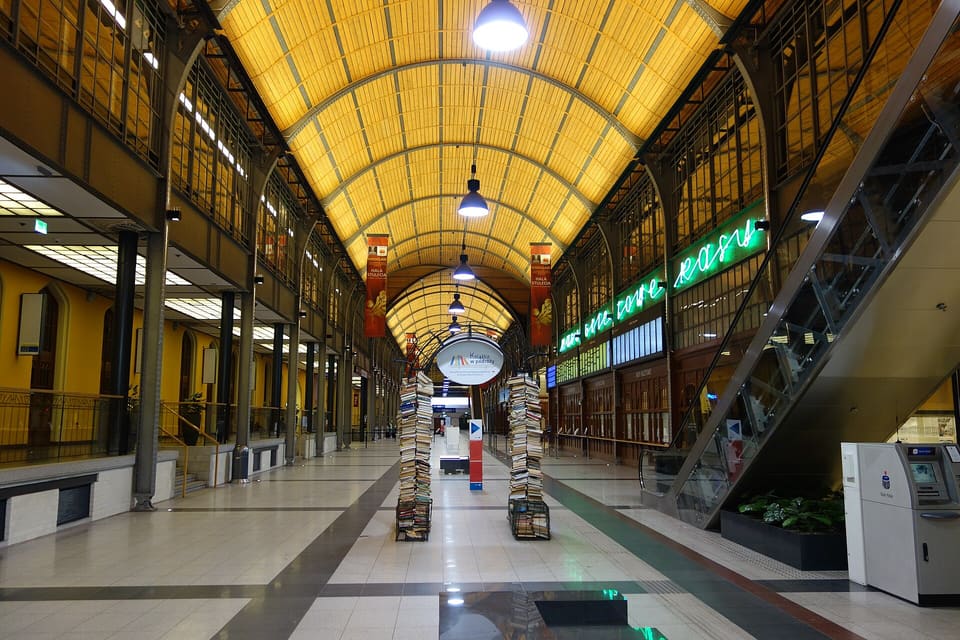 Die gewölbte Schalterhalle des Bahnhofs mit Holzdecke von oben gesehen, mit verglasten Geschäften auf beiden Seiten, Hängelampen und einer Rolltreppe im Vordergrund.