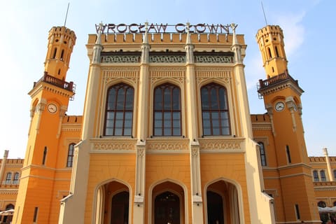 The central pavilion of Wrocław Główny station, with three arched entrances, tall arched windows, two clock towers and the station name lettered on the roof, under a pale blue sky.