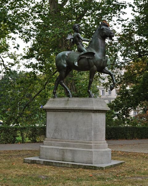 A dark bronze winged boy riding a prancing horse on a tall plain stone plinth, among trees and a hedge.