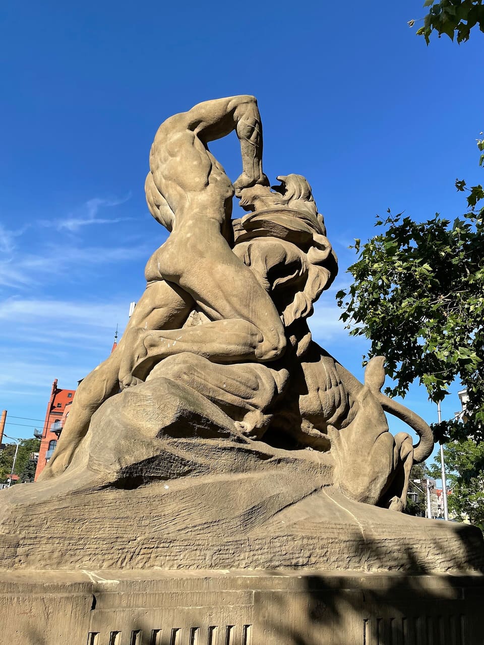 A sandstone sculpture seen from below against a blue sky: a muscular young man with one arm raised, wrestling a lion on a rocky base.