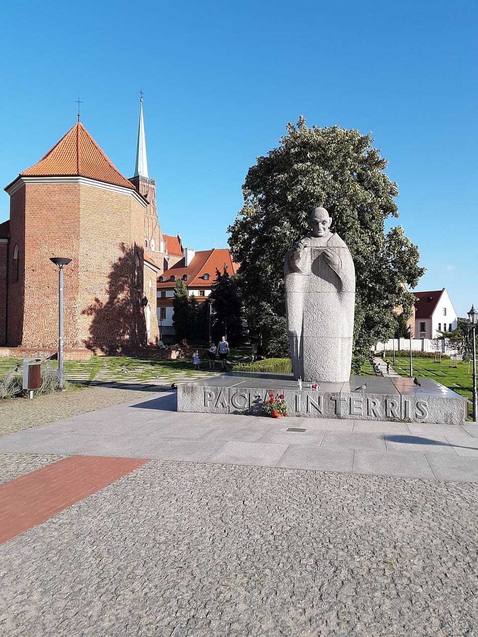 A grey granite figure of the Pope in a long robe on a low slab of stone, the words PACEM IN TERRIS cut along its front edge, with a red-brick church and a tall tree behind.