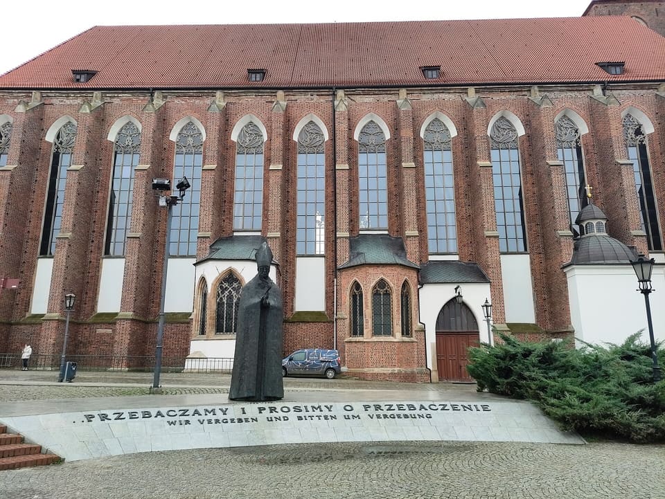 The dark bronze figure of Cardinal Kominek in a mitre and cope, standing before the long brick wall and tall windows of the Church of Our Lady on the Sand, with the Polish and German inscription cut in the pale stone ground in front of him.