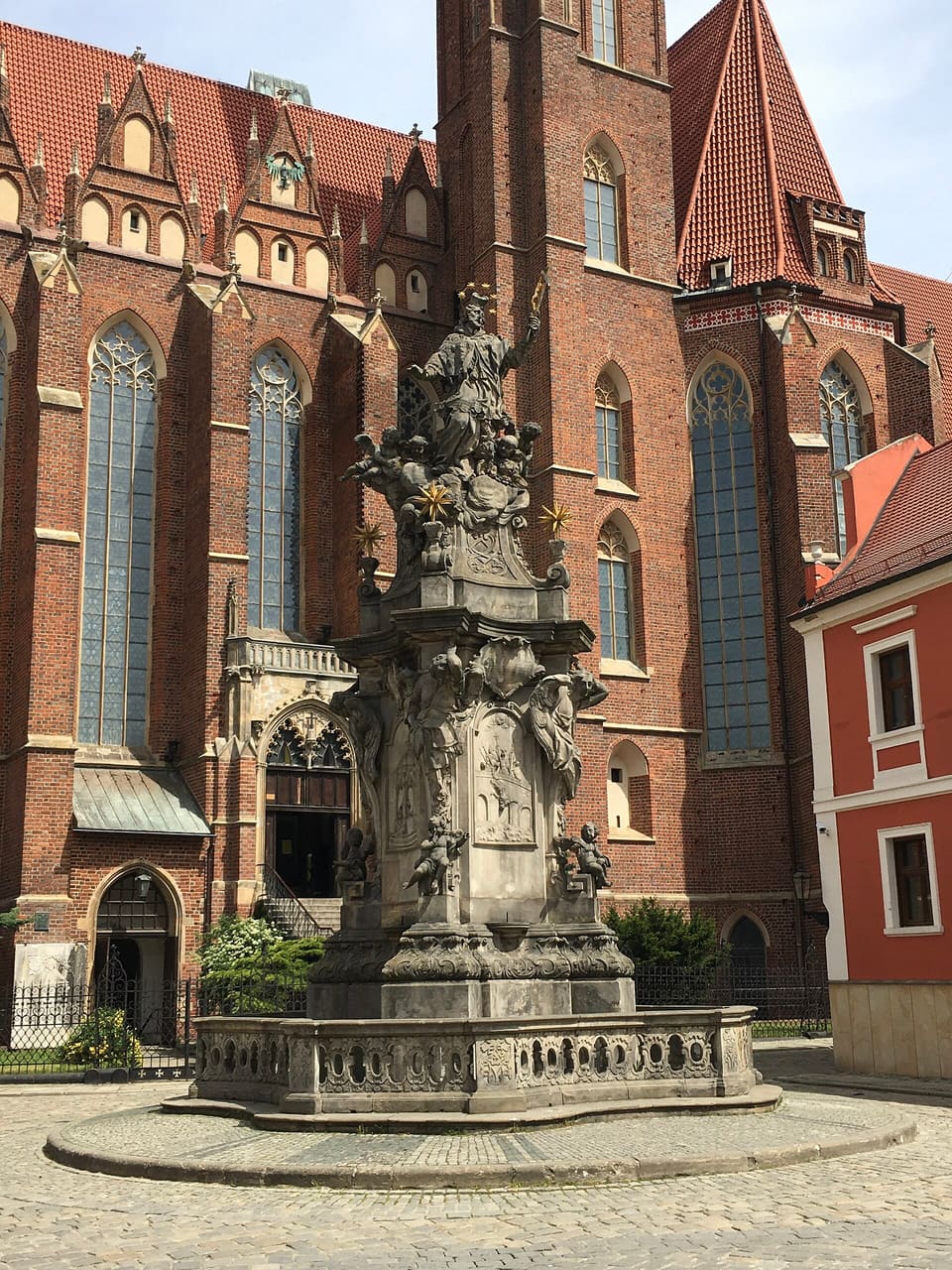 The stone monument of St John of Nepomuk on its round stepped base inside a balustrade, a saint with a raised crucifix and a ring of stars above a crowd of cherubs and angels, against the red-brick walls of the Holy Cross church.
