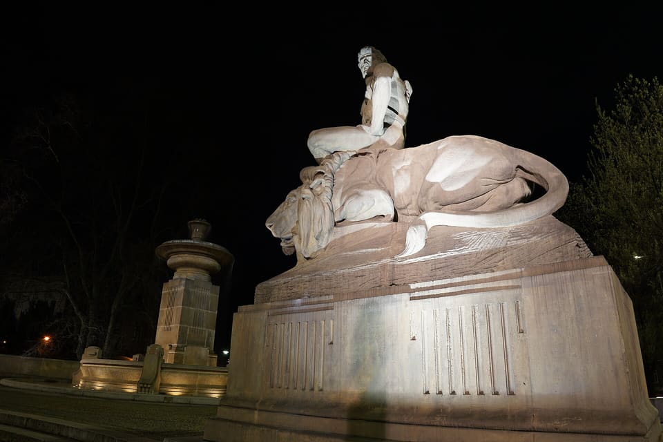 The floodlit sculpture of a man sitting on a lion at night, with the stone fountain basin and its tall central column at the left.