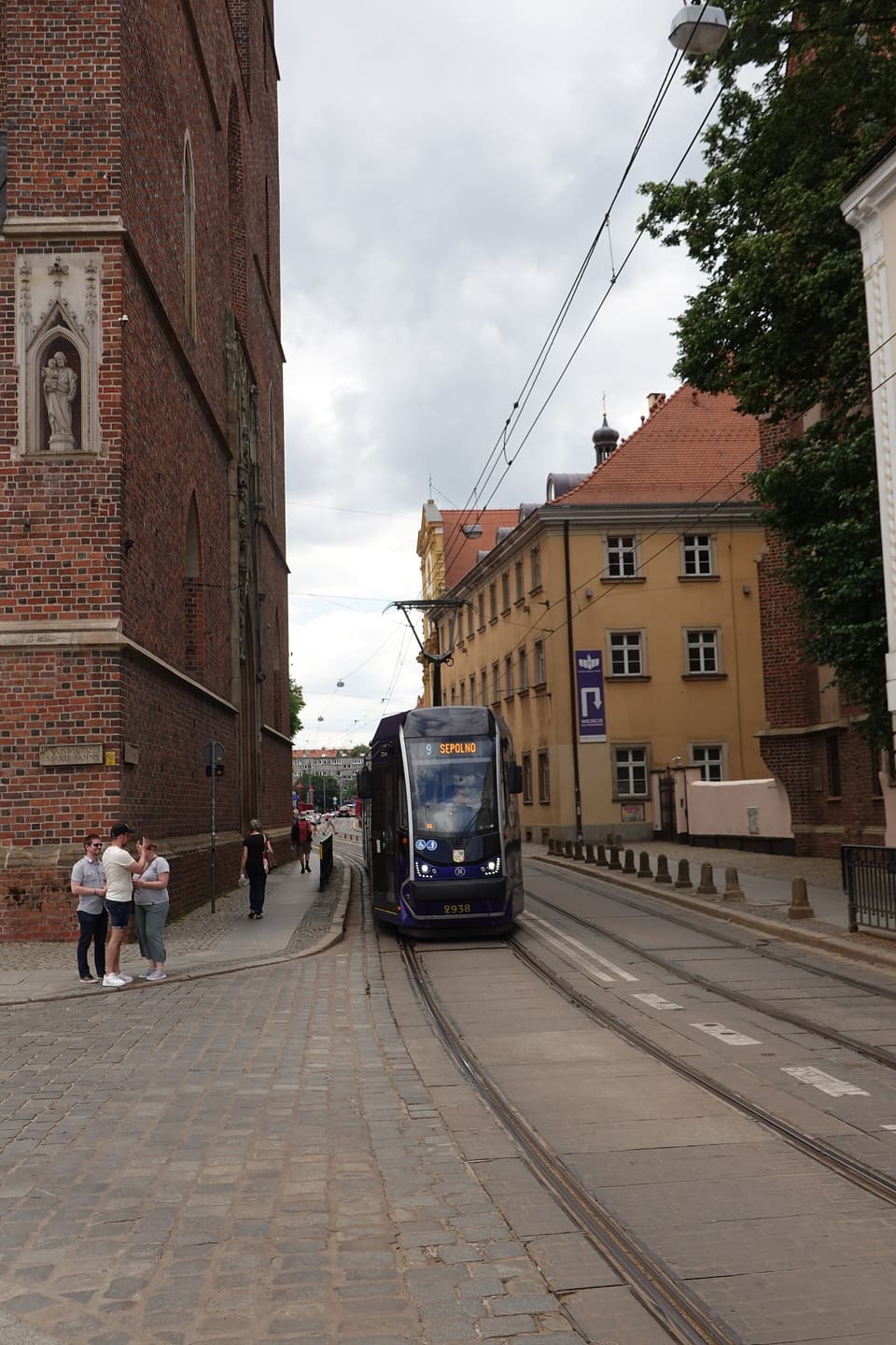 Eine lila-weiße Straßenbahn der MPK Wrocław, Linie 9 nach Sępolno, hält auf einer gepflasterten Straße im Zentrum, daneben ein paar Menschen.