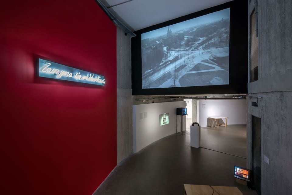 Inside the museum: bare concrete walls and pillars around a curving gallery, a line of neon handwriting on a red wall at the left, and a black-and-white film of a snowy city square projected high across an opening two storeys tall.