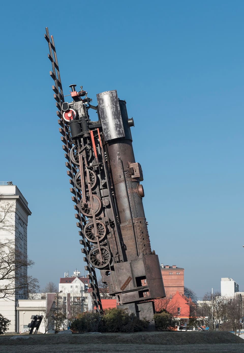 Train to Heaven: a rust-brown steam locomotive reared up almost on end along a length of track whose sleepers climb past its chimney, its wheels and rods facing the camera, against a clear blue sky, with the museum's pale front at the left edge.