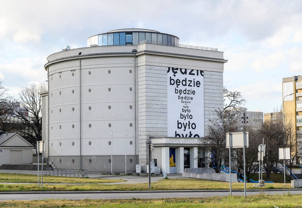 The museum seen across the road from plac Strzegomski: a pale round concrete tower ringed with rows of small round vents, a flat front block carrying the words będzie and było in large black letters, a columned porch at its foot, and a glass drum on the roof.