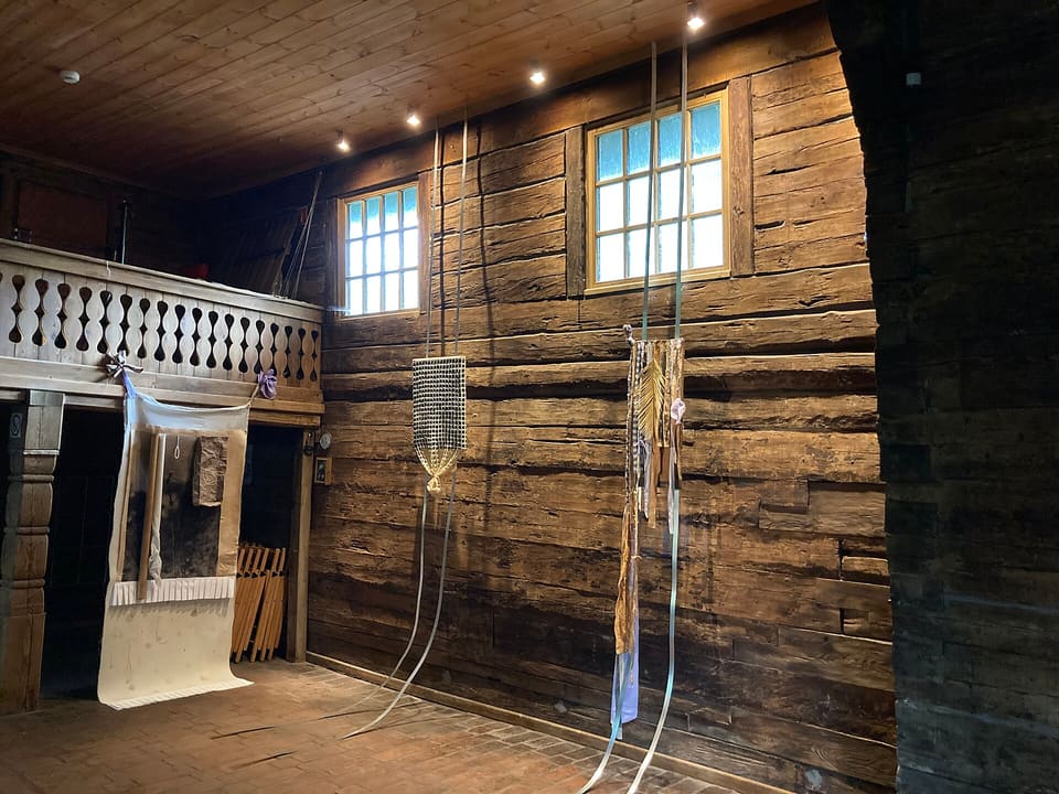 Inside the wooden church: a plank gallery along the far wall above an open arcade, with exhibition panels and hanging pieces in the room.