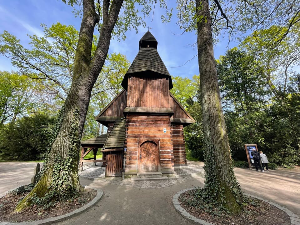 The wooden church of St John of Nepomuk seen along a path between two large tree trunks, its shingled roof and small tower rising behind them.