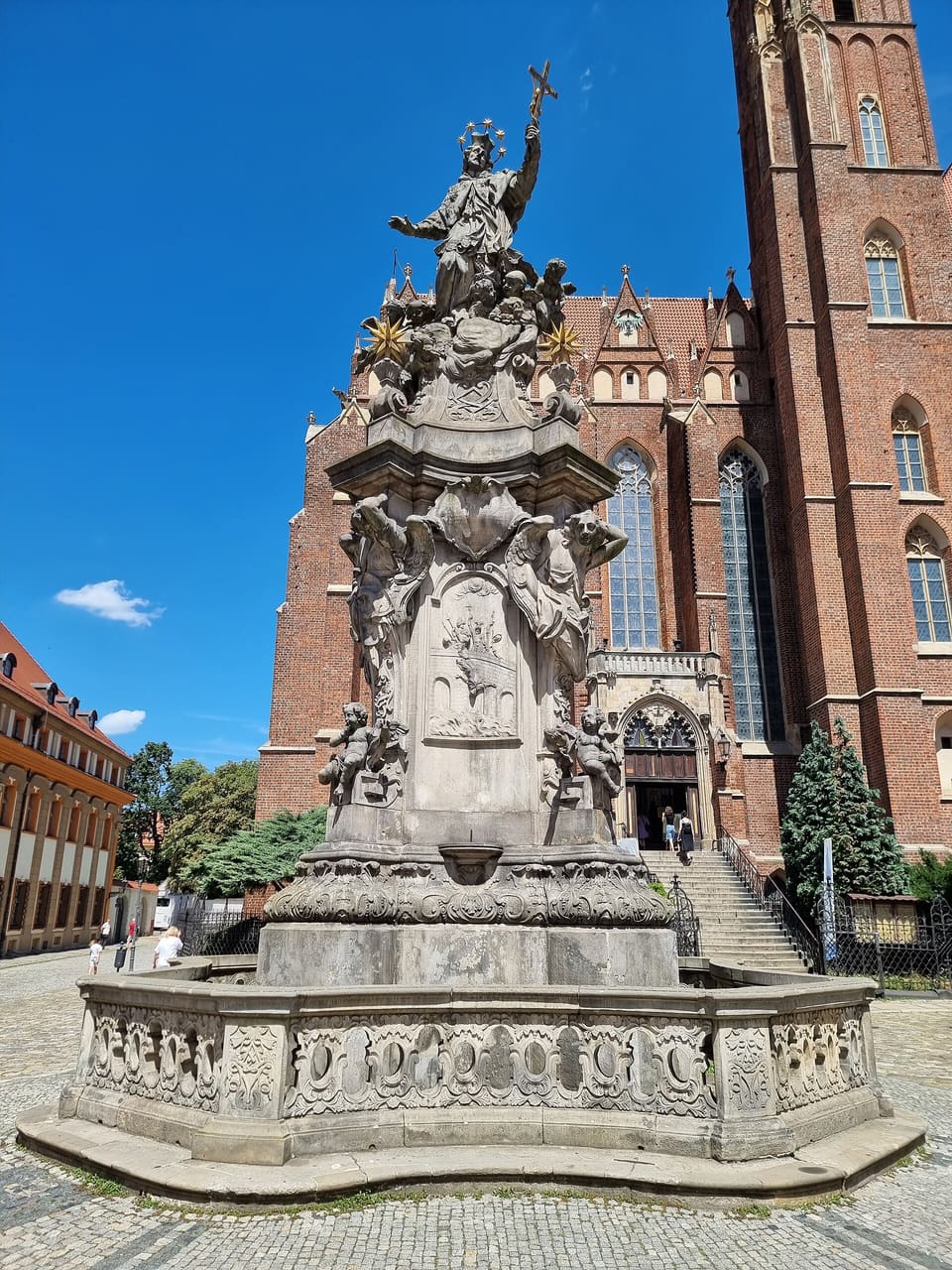 Das steinerne Denkmal des hl. Johannes Nepomuk aus wenigen Metern Entfernung: der Heilige mit erhobenem Kruzifix hoch oben auf seiner Säule aus Wolken und Putten, dahinter in der Sonne die hohe Wand aus rotem Backstein und die gotischen Fenster der Heilig-Kreuz-Kirche.